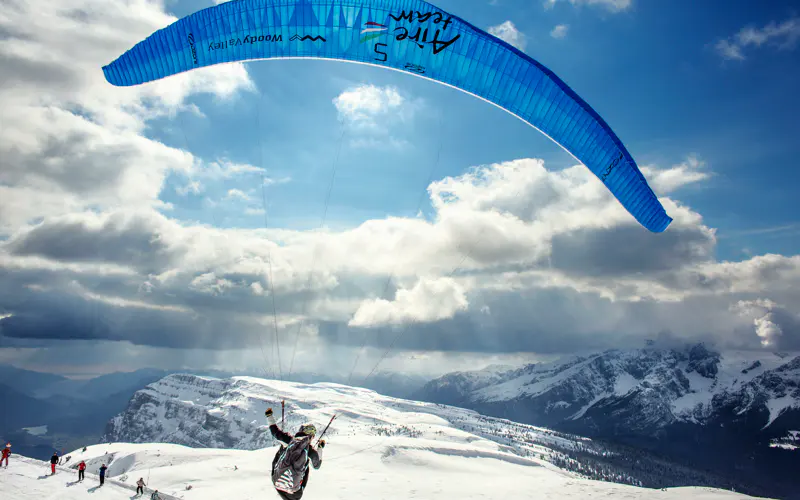 Iemand zweeft met een blauwe parachute over een besneeuwd berglandschap, terwijl een aantal anderen in de buurt wandelen of skiën onder een bewolkte hemel.
